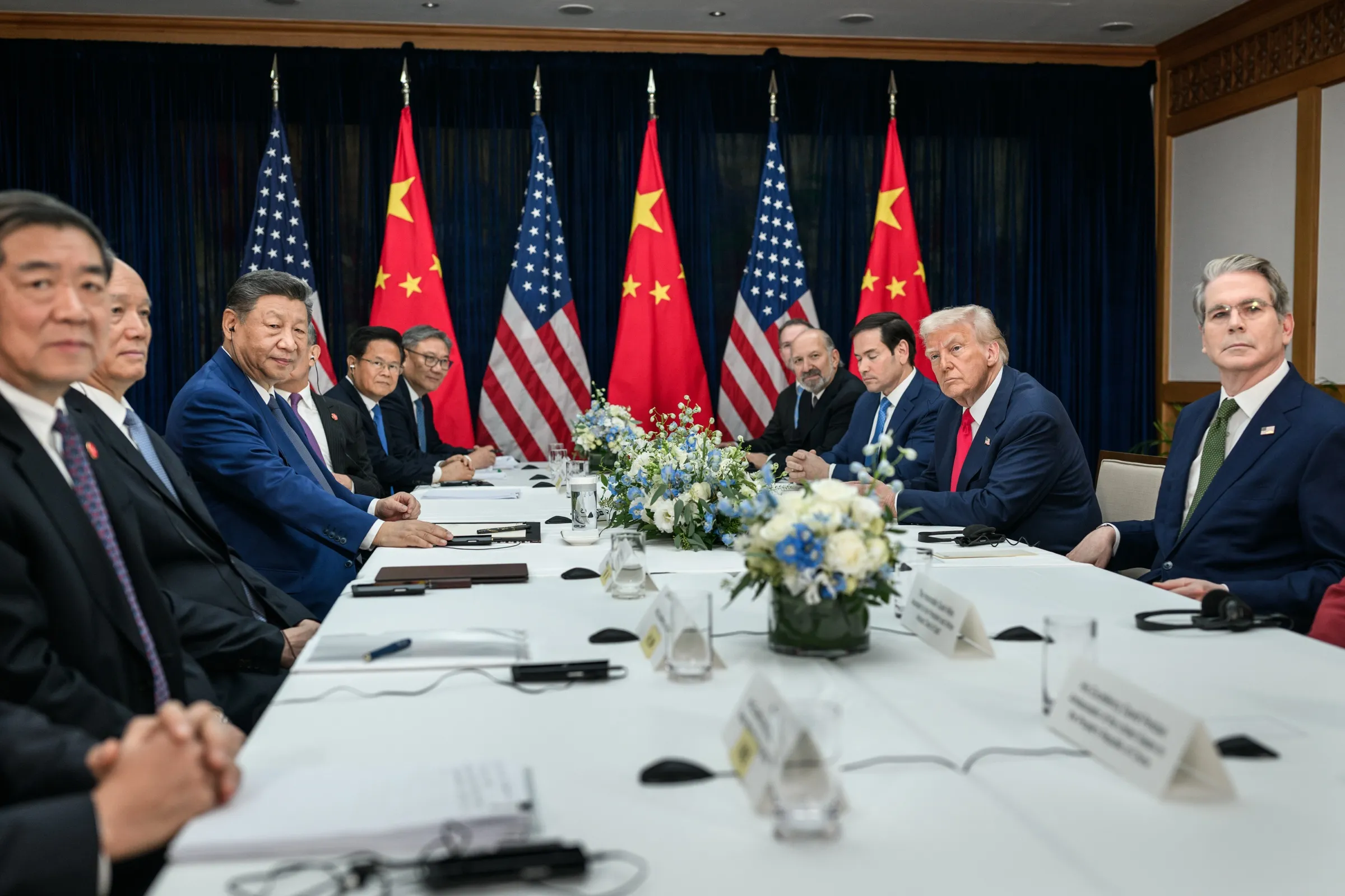 Xi Jinping and Donald Trump sit opposite one another with their delegations at a long table beneath alternating Chinese and American flags in Busan.
