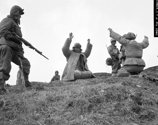 A United States Marine with a fixed bayonet stands over two kneeling Chinese prisoners who raise their hands on a low winter ridge near Hoengsong, Korea.