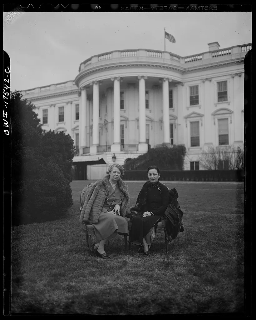 Eleanor Roosevelt and Soong Mei-ling sit side by side on chairs on the White House lawn, looking directly at the camera with the South Portico and United States flag behind them.